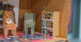 Two miniature cardboard armchairs are sat in a cardboard dollhouse. Collages pieces of colour are seen throughout.