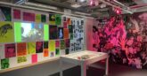 A vibrant exhibition space with colourful posters and flyers covering the walls, primarily in neon green, pink, orange, and yellow, promoting club events and music nights. A wall features a large black-and-white photo collage of club-goers in a crowded, energetic scene, partially illuminated by pink lighting. In the centre of the room is a white display table with small artifacts inside, and above it are industrial-style exposed ceilings with spotlights and pipes. A projection screen displays a video on the left wall.
