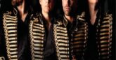 Four artists: Jonathan Ansell, Michael Christie, Duncan Sandilands, Michael Conway standing side by side, dressed in matching ornate black jackets with gold military-style trim. They are posing against a dark, textured background, looking in different directions with serious expressions.