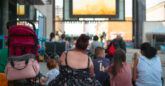 A group of people sitting on the ground outside looking at a large film screen