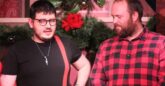 Two men stand in front of a festive holiday background with garlands, red poinsettias, and Christmas decorations. The man on the left wears glasses, a black t-shirt with a red strap across his chest, a necklace, and bracelets. The man on the right wears a red and black plaid shirt and has a beard. They appear to be engaged in conversation or part of a performance.