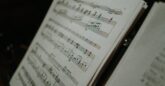 A close-up image of sheet music on a music stand, showing detailed musical notations and staffs. The sheet is angled slightly, with another page partially visible to the right.