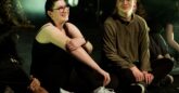 A group of young people sitting on the floor in a theatre, with stage lighting and background haze. A young girl with fair skin and dark braided hair, wearing glasses, a black sleeveless top, black leggings, and white sneakers with light blue accents is sitting cross-legged with her arms wrapped around her knees and is smiling warmly, looking slightly to her left. Next to her, on the right, is a young man with fair skin and short brown hair, also wearing glasses. He is dressed in a brown long-sleeved top and black pants, sitting cross-legged with his hands resting on his knees. He has a subtle smile and is looking toward the young girl.