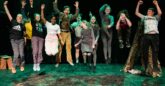 A group of young people in a row, jumping up in the air on a theatre stage. They are in costumes and have green and yellow stage lighting shining over them. They are smiling and laughing.