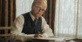 A man sits by a large window at a desk with papers on, writing