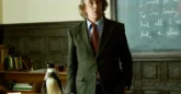 A man wearing a jacket and tie stands before a blackboard. On a desk next to him is a penguin