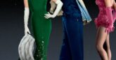 A man in a blue suit stands between a woman in a long green dress and a woman in a pink showgirl outfit