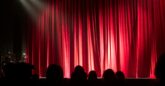 A theatre stage with closed red curtains illuminated by stage lights, with the silhouettes of an audience seated in the foreground